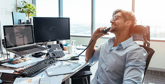 Ein Mann telefoniert am Büro während er dabei lächelt und auf einem Bürostuhl sitzt Ein Mann telefoniert am Büro während er dabei lächelt und auf einem Bürostuhl sitzt