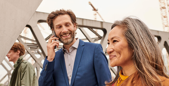 Geschäftliches Gespräch auf der Brücke Mann im Anzug telefoniert, Kollegin lächelt neben ihm auf einer modernen Brücke