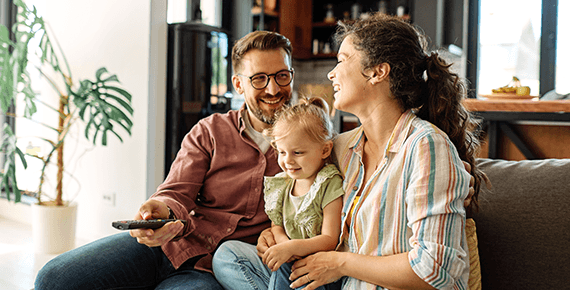 Eine Familie sitzt glücklich lachend auf einem Sofa im Wohnzimmer. Der Mann hält eine Fernbedienung.