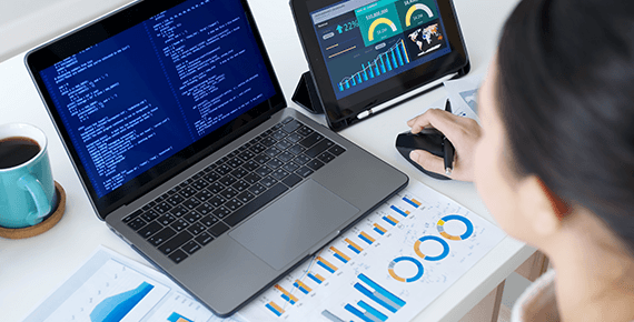Ein Laptop Bildschirm zeigt Codezeilen. Am Schreibtisch liegt um den Laptop herum ausgedruckte Grafiken und Diagramme