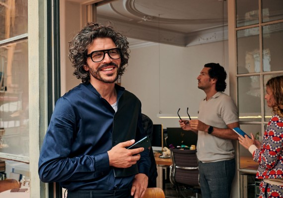 Mann lächelt mit Smartphone am Büroeingang, während Kollegen im Hintergrund arbeiten