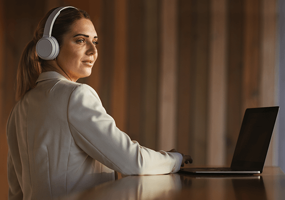 Eine junge Frau lehnt an einem Tisch auf dem ein Laptop steht. Dabei trägt sie Kopfhörer