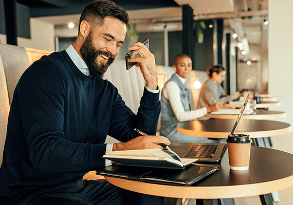 Ein Mann in blauem Pullover sitzt in einem Cafe, telefoniert und macht sich dabei Notizen