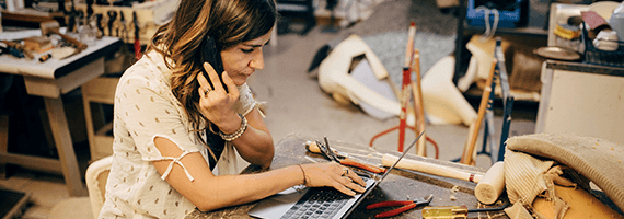 Eine Frau sitzt in einer Werkstatt am Laptop und telefoniert dabei