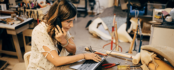 Eine Frau sitzt in einer Werkstatt am Laptop und telefoniert dabei