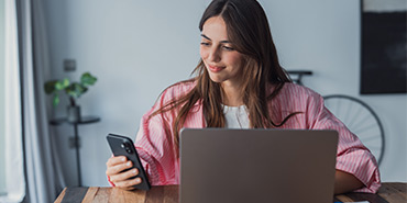 Eine Frau sitzt am Tisch mit ihrem Laptop und einem Smartphone in der Hand.