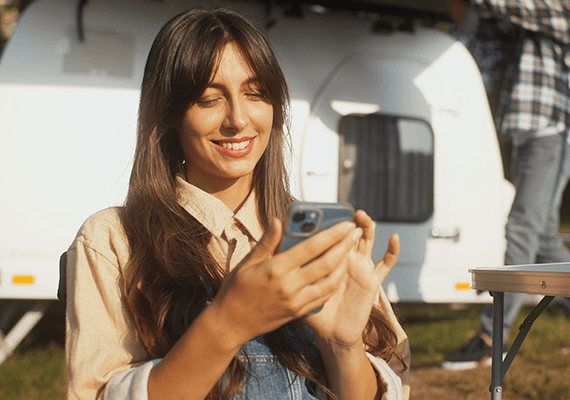 Eine Frau hält ihr Mobiltelefon. Im Hintergrund ist unscharf ein Wohnwagen zu sehen.