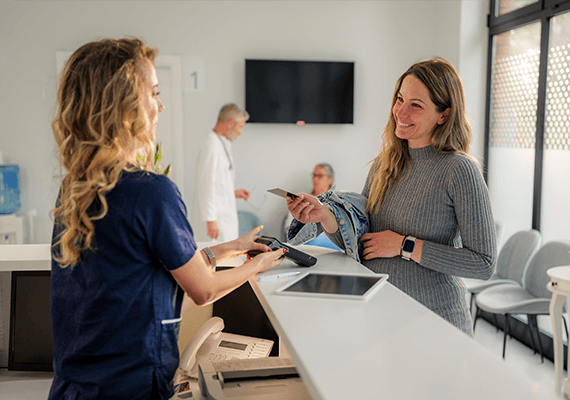 Eine Frau steht lächelnd in einer Arztpraxis einer Dame in blauer Dienstuniform gegenüber. Sie hält ihre Bankomatkarte hoch. Die Frau gegenüber hält ihr ein Kartenlesegerät entgegen.