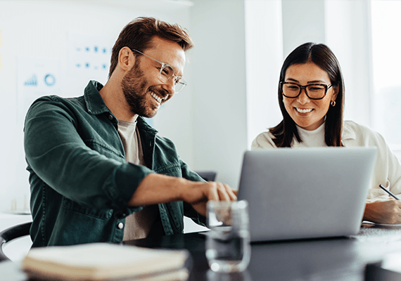 Ein Mann und eine Frau unterhalten sich an einem Schreibtisch in einem hellen Büro. Sie lächeln dabei und der Mann zeigt auf einen Laptopbildschirm.