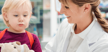 Frau in weißem Kittel lächelt ein Kind mit Kuscheltier in einer Apotheke an.
