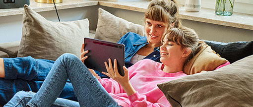 Zwei junge Frauen liegen auf einem Sofa und schauen in ein Tablet