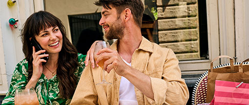 Junges Paar sitzt an einem Tisch in einem Café im Freien und genießt Getränke. Die Frau telefoniert lächelnd. Der Mann hält ein Glas mit einem Getränk, während er sie anblickt. 
