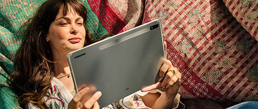 Junge Frau mit braunen, welligen Haaren liegt entspannt in einer Hängematte und hält ein silbernes Tablet in den Händen.