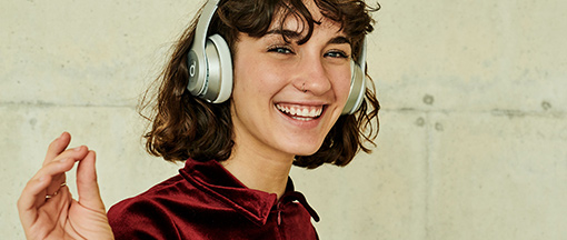 Frau mit braunen Haaren trägt silberne Kopfhörer und macht eine entspannte Handgeste vor heller Wand