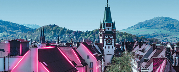 Blick über ein Altstadtdach mit pinken Lichtlinien, im Hintergrund ein Uhrturm und grüne Hügel unter klarem Himmel.