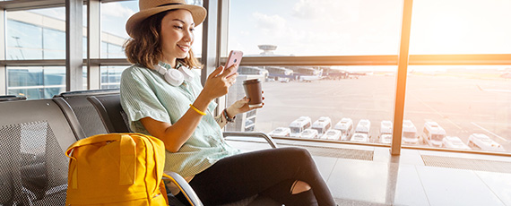 Frau am Flughafen Eine junge Frau mit Hut und lockigen Haaren sitzt in einem Wartebereich am Flughafen. Sie trägt Kopfhörer um den Hals, hält ein Smartphone in der einen und einen Kaffeebecher in der anderen Hand.