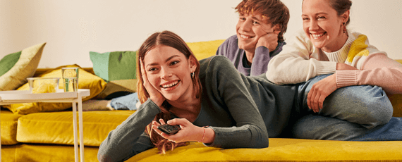 Junge Menschen genießen gemeinsam einen Fernsehabend Drei junge Erwachsene liegen entspannt auf einem Sofa und lachen, eine Frau hält eine Fernbedienung
