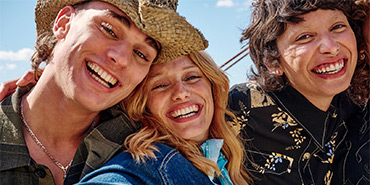 Drei fröhliche junge Menschen umarmen sich draußen bei Sonnenschein, lachen in die Kamera, einer trägt einen Strohhut.