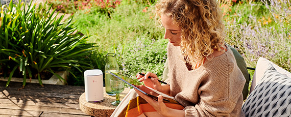 Frau sitzt entspannt im Garten, arbeitet mit Tablet und Stylus, daneben ein Getränk und ein kompakter weißer Router auf einem Tisch.