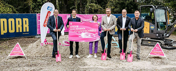 Sechs Personen mit pinken Spaten feiern Spatenstich für Glasfaserausbau in Steyr, vor Baustelle mit Bagger und Werbebannern.