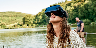 Frau erlebt virtuelle Realität mit VR-Brille am See – moderne Technik draußen genießen dank leistungsstarker Internetverbindung.