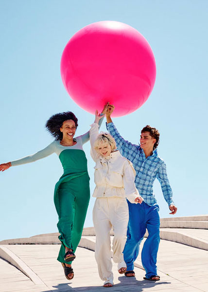 Entertainment Drei fröhliche Menschen laufen Hand in Hand im Sonnenschein, halten gemeinsam einen riesigen pinken Ballon unter blauem Himmel.