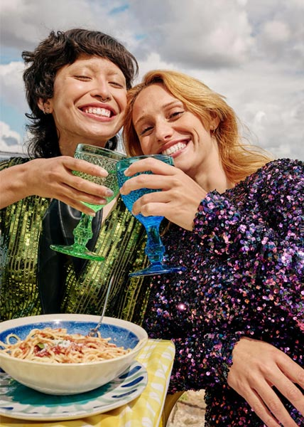 Essen und Trinken Zwei lachende Frauen stoßen mit bunten Gläsern an, sitzen an einem Tisch mit Pasta unter freiem Himmel und genießen die gemeinsame Zeit.