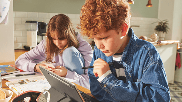 Zwei Kinder sitzen mit Tablet und Handy in der Küche am Tisch.