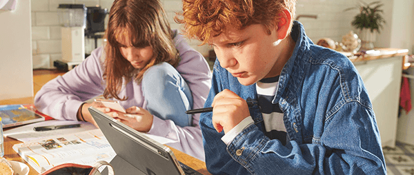 Zwei Kinder sitzen mit Tablet und Handy in der Küche am Tisch.