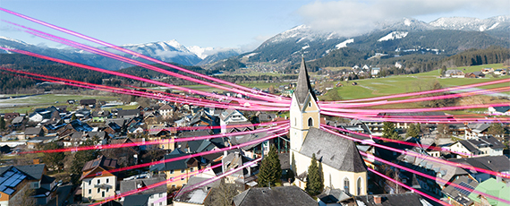 Luftaufnahme eines Dorfs in den Alpen, pinke Linien symbolisieren digitale Glasfaservernetzung, ausgehend von einer Kirche im Zentrum.