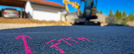 Pinkes „FTTH“-Markierungsspray mit Pfeil auf Asphalt, im Hintergrund ein Bagger und ein Haus – Hinweis auf Glasfaserverlegung bis ins Haus.