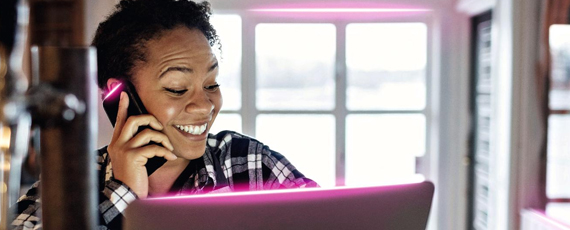 Lächelnde Frau telefoniert und arbeitet gleichzeitig am Laptop, magentafarbene Lichtakzente betonen digitale Vernetzung im Homeoffice.