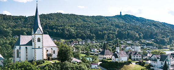 Maria Wörth Panoramaaufnahme von Maria Wörth mit Kirche, umgeben von grünen Hügeln und Wald, im Hintergrund ein Aussichtsturm auf dem Berg.