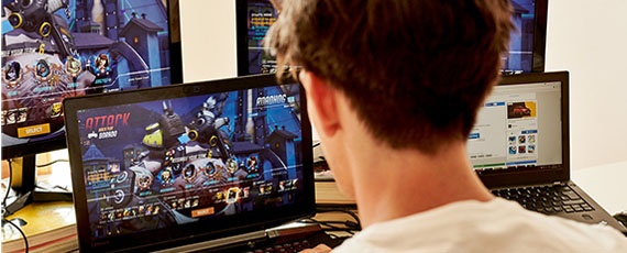 Junger Mann sitzt vor mehreren Bildschirmen und spielt ein Online-Game auf dem Laptop, im Hintergrund laufen weitere Gaming-Sessions.