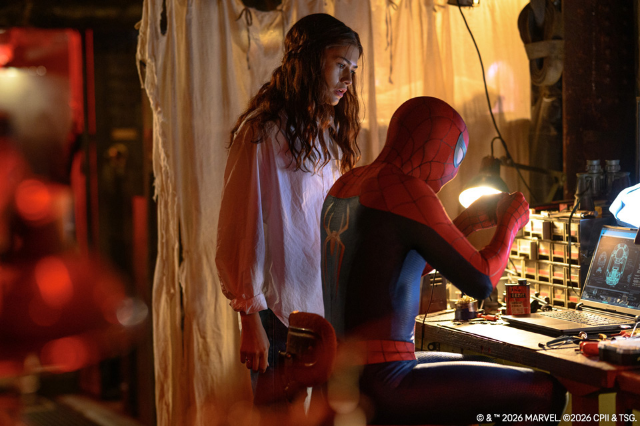 .Spider-Man sitzt an einem technischen Arbeitsplatz, während eine Frau neben ihm steht und zusieht.