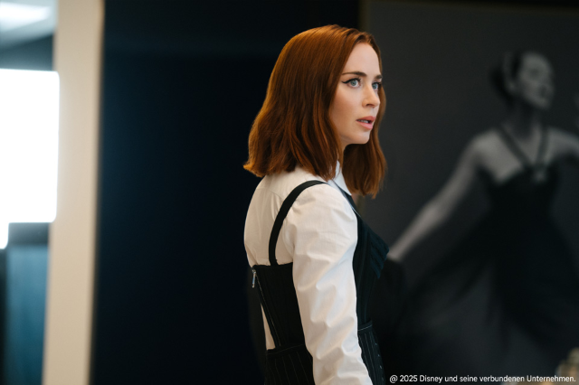 Eine rothaarige Frau trägt eine weiße Bluse unter einem schwarzen Kleid und blickt aufmerksam zur Seite in einem modernen, stilvollen Innenraum.